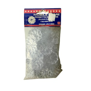 Op de afbeelding: Een doorzichtige plastic zak met doorzichtige rozetknoppen. De zak heeft een blauw en rood label met de tekst "MOBILE HOME ROSETTE BUTTONS" en "100 PER BAG". De rozetknoppen hebben een diameter van ongeveer 2,8 cm.