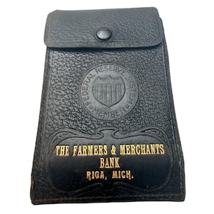 以下が含まれることがあります： スナップ留めの黒い革製財布。前面には、"THE FARMERS & MERCHANTS BANK RIGA, MICH."のテキストと連邦準備制度のエンブレムが金色のエンボス加工で施されています。
