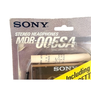 May include: A Sony MDR-005SA stereo headphones package with a grey cardboard backing. The clear plastic blister pack contains the headphones and a white cylindrical component with text. A yellow sticker on the bottom right reads Including.