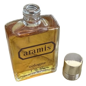 May include: A vintage Aramis cologne bottle with a gold-colored liquid inside. The bottle is rectangular with a clear glass top and a gold cap. The label reads "aramis cologne 1 FL. OZ."