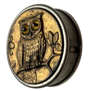 May include: A round, antique-style metal button with an embossed owl design. The owl has yellow eyes and is perched on a branch with leaves. The button has a gold and silver color scheme.