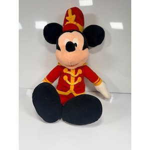 May include: A plush Mickey Mouse doll wearing a red and yellow band uniform. The doll has black ears, a red hat, and black shoes. The doll is sitting with its legs extended.