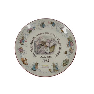 Könnte beinhalten: Ein runder Keramikteller mit einer zentralen Illustration von Mrs. Tiggy-Winkle, umgeben von kleinen Tierfiguren, die Bücher halten. Der Text lautet Mrs. Tiggy-Winkle wishes you a very Happy Birthday, Beatrix Potter, 1982, mit einem dünnen roten Rand.