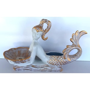 May include: A white and gold ceramic mermaid figurine holding a scalloped shell bowl. The mermaid has a high ponytail, a detailed tail with gold-painted scales, and a base textured to resemble waves, all accented with a distressed gold finish.
