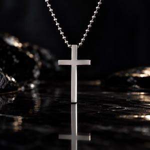 May include: A polished silver-toned cross pendant suspended from a matching silver-toned ball chain. The cross features a smooth, flat surface with clean, straight edges, reflecting light against a dark, reflective surface below the pendant.