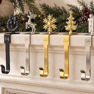 May include: Six metal stocking holders with personalised names engraved on the vertical shafts. The hooks feature decorative toppers including a black wreath, silver reindeer, gold snowflake, gold Christmas tree, silver gift box, and black bell.