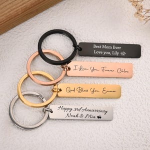 Puede incluir: Una colección de llaveros personalizados con mensajes grabados. Los llaveros presentan varios acabados metálicos, incluyendo negro, oro rosa, oro y plata. Cada llavero tiene un anillo y una etiqueta rectangular con texto como "Best Mom Ever" e "I Love You Forever."
