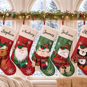Peut inclure: Cinq chaussettes de Noël avec des revers blancs, personnalisées avec les prénoms Sophia, Audrey, James, Julian et Stella. Elles présentent des personnages 3D en peluche : renne, père Noël, elfe, bonhomme de neige et pingouin, portant des écharpes.