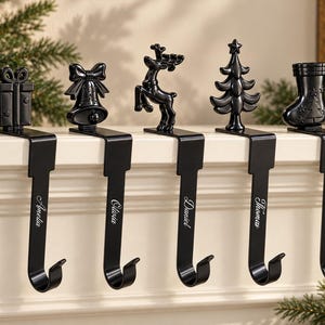 May include: Five black metal stocking holders with unique holiday-themed toppers including a gift, bell, reindeer, tree, and stocking. Each hook features a personalized name engraved in white script: Amelia, Olivia, Daniel, Thomas, and Jayden.