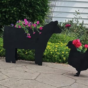 May include: Three black metal animal-shaped planters on a stone patio. The planters are shaped like a cow and two chickens. Each planter is filled with colourful flowers, creating a decorative garden display.