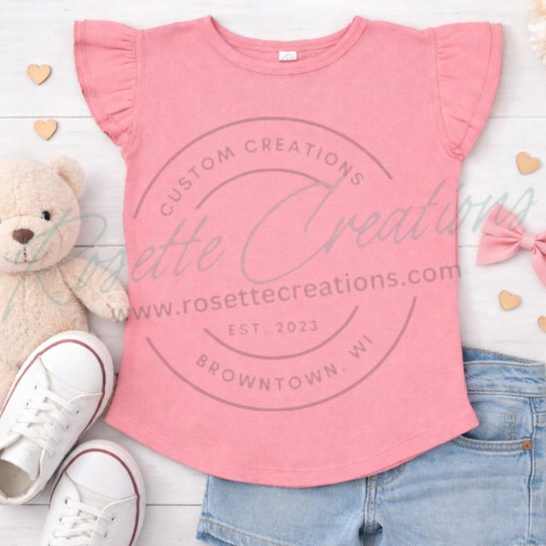 May include: A pink t-shirt with ruffled sleeves, featuring the text "Custom Creations" and the website address rosetteecreations.com. The shirt is paired with denim shorts, white sneakers, a teddy bear, and a pink bow. The text also includes "EST. 2023" and "Browntown, WI."