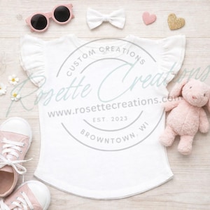 May include: A white ruffle-sleeve t-shirt with the text "CUSTOM CREATIONS" and "www.rosettecreations.com" is displayed on a light wood surface. Accessories include pink sunglasses, a white bow, a pink heart, a gold heart, a pink stuffed bunny, and pink sneakers.
