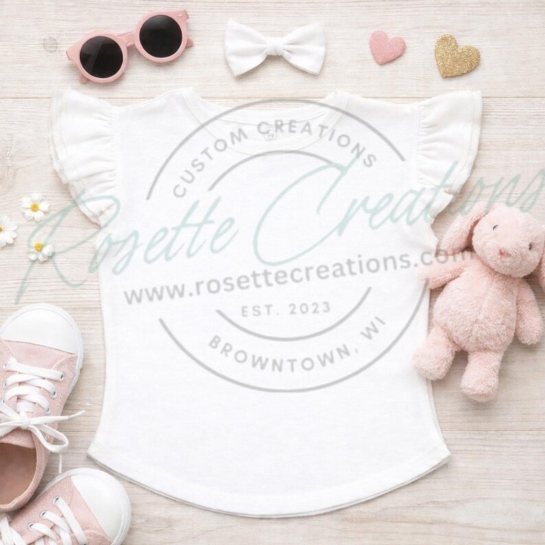 May include: A white ruffle-sleeve t-shirt with the text "CUSTOM CREATIONS" and "www.rosettecreations.com" is displayed on a light wood surface. Accessories include pink sunglasses, a white bow, a pink heart, a gold heart, a pink stuffed bunny, and pink sneakers.