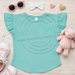 May include: A turquoise ruffle-sleeve t-shirt with the text "Rosette Creations" and a pink stuffed bunny, sunglasses, and shoes. The shirt is displayed on a light wood surface with a white bow and heart-shaped decorations.