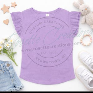 May include: A lilac-colored t-shirt with ruffled sleeves. The shirt has a round neck and a curved hem. The image includes a pair of white sneakers, denim shorts, a stuffed bunny, and wooden stars and hearts.