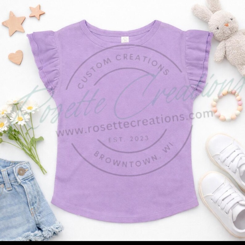 May include: A lilac-colored t-shirt with ruffled sleeves. The shirt has a round neck and a curved hem. The image includes a pair of white sneakers, denim shorts, a stuffed bunny, and wooden stars and hearts.