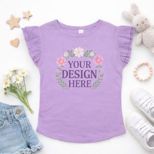 May include: A lilac-colored t-shirt with ruffled sleeves and the text "YOUR DESIGN HERE" inside a floral wreath. The shirt is surrounded by a pair of white sneakers, a stuffed bunny, and wooden shapes.