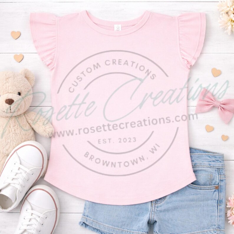 May include: A light pink t-shirt with ruffled sleeves. The shirt has a curved hem and a circular logo that reads "Custom Creations Rosette Creations" with a website address and "Est. 2023 Browntown, WI". The shirt is paired with denim shorts and white sneakers.