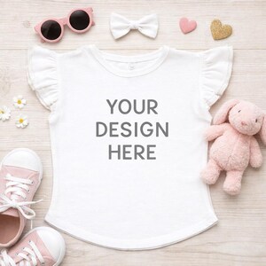 May include: White t-shirt with ruffled sleeves and the text "YOUR DESIGN HERE". The image includes pink sunglasses, a white bow, pink sneakers, a pink stuffed bunny, and decorative hearts. The items are arranged on a light wood surface.