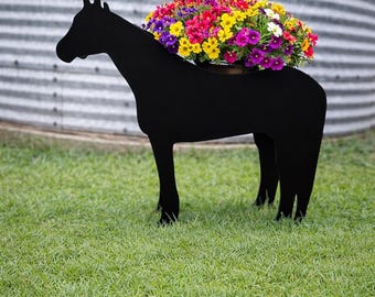 May include: A matte black metal horse-shaped planter silhouette standing on four legs. The back of the horse features a circular opening filled with a dense arrangement of vibrant, multi-colored flowers including shades of yellow, pink, and purple.