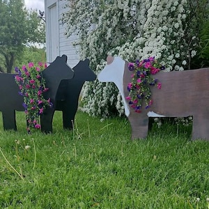 May include: Three wooden cow-shaped planters. Two are black, and one is brown with white accents. Each planter is filled with vibrant pink and purple flowers, creating a charming garden display. The planters are placed on a grassy lawn.