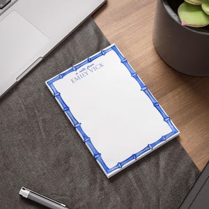 Puede incluir: Bloc de notas blanco con un borde azul estilo bambú. El bloc de notas tiene el texto azul "a note from EMILY VICK". El bloc de notas está sobre un escritorio de madera junto a un portátil y un bolígrafo.