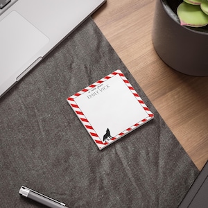 May include: Square notepad with a white interior and a red and white striped border. The notepad features the text "a note from EMILY VICK" and a black silhouette of a howling wolf. The notepad is on a gray surface.
