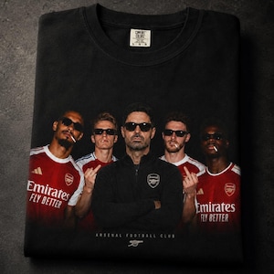 Può includere: Felpa nera girocollo con grafica di cinque persone con occhiali da sole. Le persone indossano maglie da calcio rosse e bianche con la scritta "Emirates Fly Better". Il testo "Arsenal Football Club" è stampato sotto.