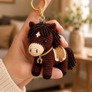 May include: A dark brown crocheted horse keychain featuring a cream-colored muzzle, a white star on the forehead, a tan saddle with a small brown button, and a gold-toned horseshoe charm hanging from a braided gold cord on a metal ring.