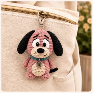 May include: A crocheted dog keychain featuring a pink body, black ears, and a cream-colored belly. The dog has large white eyes with black pupils, a black nose, and a teal collar with a small silver house-shaped charm attached to a metal clasp.