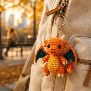 May include: A crocheted orange dragon keychain with a yellow belly, dark teal wings, and a flame-tipped tail. The small stuffed creature features two small horns, black bead eyes, and light-colored claws, attached to a silver metal keyring.