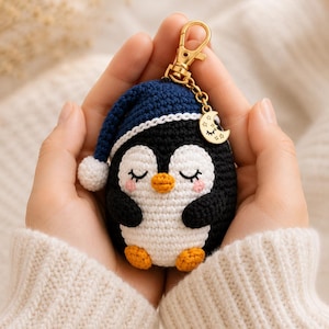 May include: A crocheted penguin keyring featuring a sleeping expression with closed eyes, a small orange beak, and pink cheeks. The penguin wears a dark blue nightcap with a white pom-pom and includes a gold-toned metal clasp and a crescent moon charm.