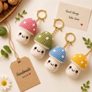 May include: Four crochet mushroom keychains in pink, green, blue, and yellow, each with a white stem, black eyes, and pink cheeks. Each keychain has a gold metal ring. A card reads Good things take time and a tag reads Handmade with love.