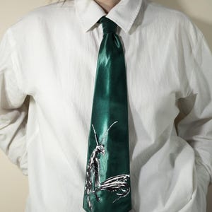 May include: A dark emerald green satin necktie featuring a detailed white line art illustration of a praying mantis positioned near the bottom. The tie has a smooth, reflective finish and is worn with a crisp, plain white button-down shirt.