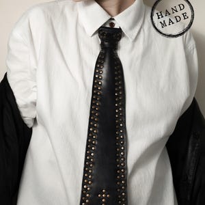 Puede incluir: Corbata de cuero negro con tachuelas de color latón. La corbata presenta un diseño de cruz en la parte inferior y se lleva sobre una camisa blanca. Un sello "HAND MADE" es visible.