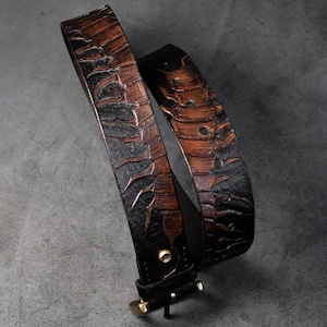 May include: A dark brown and black belt featuring a textured, embossed pattern across the strap. The belt includes a metallic buckle, a single belt loop, and a series of circular holes for adjustment, showcasing a two-tone finish throughout.