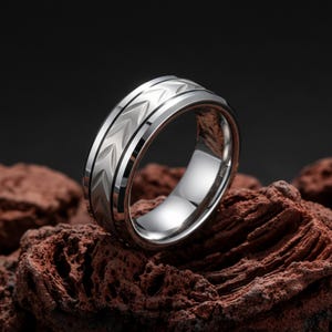 May include: A silver-colored ring with a chevron pattern. The ring has a polished finish and a band with a textured design. The ring is displayed on a reddish-brown, textured surface.