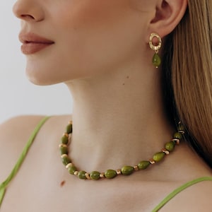 Puede incluir: Una mujer lleva un collar de cuentas verdes y pendientes a juego. El collar presenta cuentas de color verde oliva con detalles dorados. Los pendientes tienen un diseño similar, con un aro dorado y una cuenta verde colgante. Lleva un top verde.