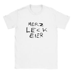 May include: White t-shirt with the black text "MERZ LECK EIER" printed on the front. The text is in a handwritten style. The shirt has short sleeves and a crew neck.
