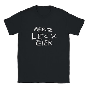 Könnte beinhalten: Schwarzes T-Shirt mit dem weißen Text "MERZ LECK EIER" im handschriftlichen Stil. Das Shirt hat einen Rundhalsausschnitt und ist für den Alltag geeignet.