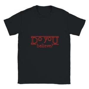Op de afbeelding: Een zwart t-shirt met korte mouwen en de tekst "DO YOU believe?" in het rood gedrukt. De belettering is gestileerd met horizontale lijnen boven en onder het woord "DO YOU" en is gecentreerd op de borst van het kledingstuk.