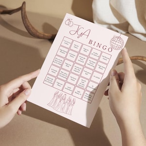 Könnte beinhalten: Eine rosa JGA Bingo-Spielkarte mit einer Grafik von Eheringen und dem Wort "BINGO" in fetter Schrift. Die Karte zeigt ein Raster von Sätzen und eine Grafik einer Discokugel. Die Karte wird von einer Person gehalten.