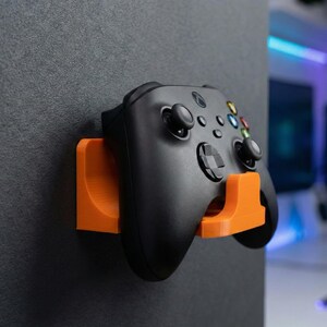 Peut inclure: Un contrôleur de jeu noir est monté sur un support mural orange. Le contrôleur comporte plusieurs boutons colorés et un pavé directionnel. Le support est fixé à un mur gris foncé, offrant un rangement peu encombrant pour les accessoires de jeu.