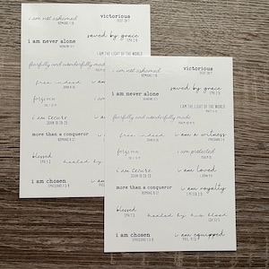 May include: Two white rectangular stickers with black handwritten text. The stickers feature phrases like "I am not ashamed," "Victorious," and "Saved by Grace," along with corresponding Bible verses. The stickers are on a wooden surface.
