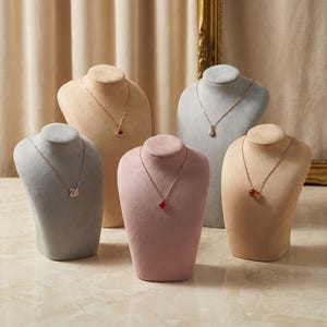May include: Five necklace displays with various pendant necklaces. The displays are in shades of beige, gray, and pink. The necklaces feature pendants in the shapes of a swan, a clover, and other gemstone designs. The background is a neutral beige.