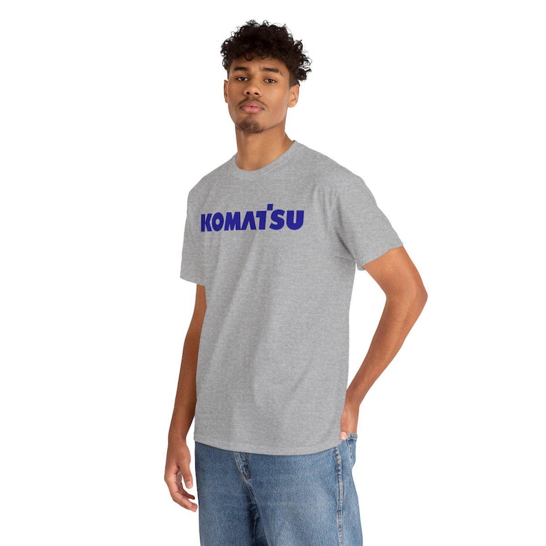 May include: A light gray t-shirt with the word "KOMATSU" printed in blue across the chest. The shirt is made of a soft material and has short sleeves. The model is wearing blue jeans.