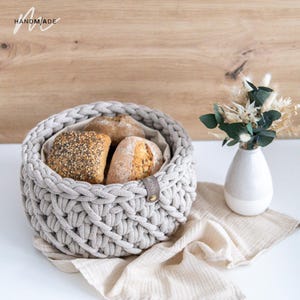 Könnte beinhalten: Ein handgefertigter, hellgrauer, gewebter Korb gefüllt mit verschiedenen Brötchen. Der Korb hat ein geflochtenes Design und ein Lederetikett. Das Wort "HANDMADE" befindet sich in der oberen linken Ecke. Rechts befindet sich eine kleine weiße Vase mit Trockenblumen.