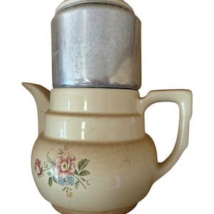May include: Vintage cream-colored coffee pot with a floral design. The pot has a silver metal filter and lid. The floral pattern includes pink, blue, and yellow flowers with green leaves. The pot has a handle and a spout.