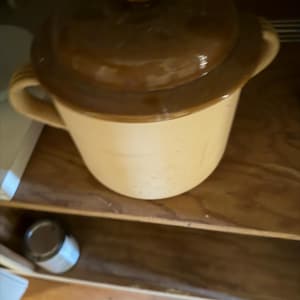 Peut inclure: Un pot en céramique beige et marron avec un couvercle. Le pot a deux poignées et un couvercle avec un bouton. Le pot est posé sur une étagère en bois. Une petite boîte est visible en dessous.