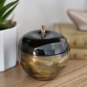May include: An apple-shaped trinket box with a glossy black top and a gold and bronze swirling pattern on the lower half. It features a small, textured stem. The box is a decorative item.